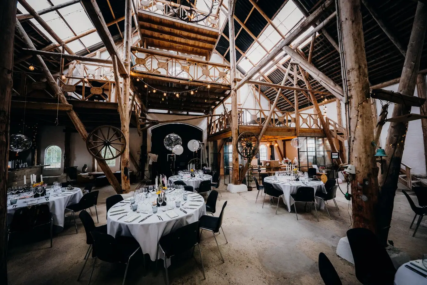Grande grange en charpente de bois, tables rondes dressées et guirlandes lumineuses
