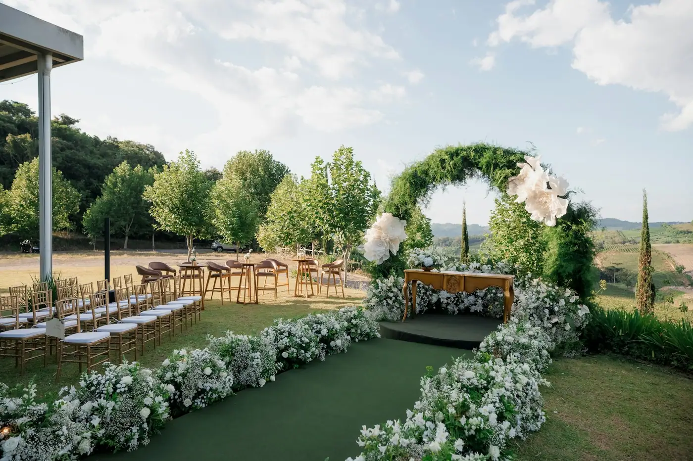 Arche de verdure et de fleurs blanches au bout d’une allée, rangées de chaises face aux collines